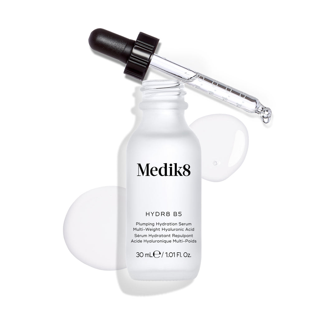 La botella del sérum Medik8 Hydr8 B5 se encuentra sobre un fondo blanco limpio con un charco de líquido transparente y gotas, mientras el cuentagotas descansa en la parte superior. El texto identifica el producto como un “Sérum líquido rehidratante” de 30 mL.