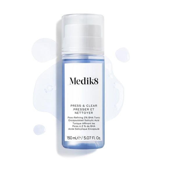 Botella de Medik8 Press & Clear (150 ml) mostrada sobre un fondo blanco con salpicaduras de tónico azul a su alrededor. El texto destaca la fórmula de tónico exfoliante con 2 % de BHA.-17