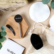 Una composición en flat lay de accesorios ecológicos de Medik8 sobre un fondo beige, incluyendo un exprimidor metálico de tubos, una espátula metálica, discos de algodón reutilizables de bambú, una bolsa de malla y una esponja de maquillaje negra.