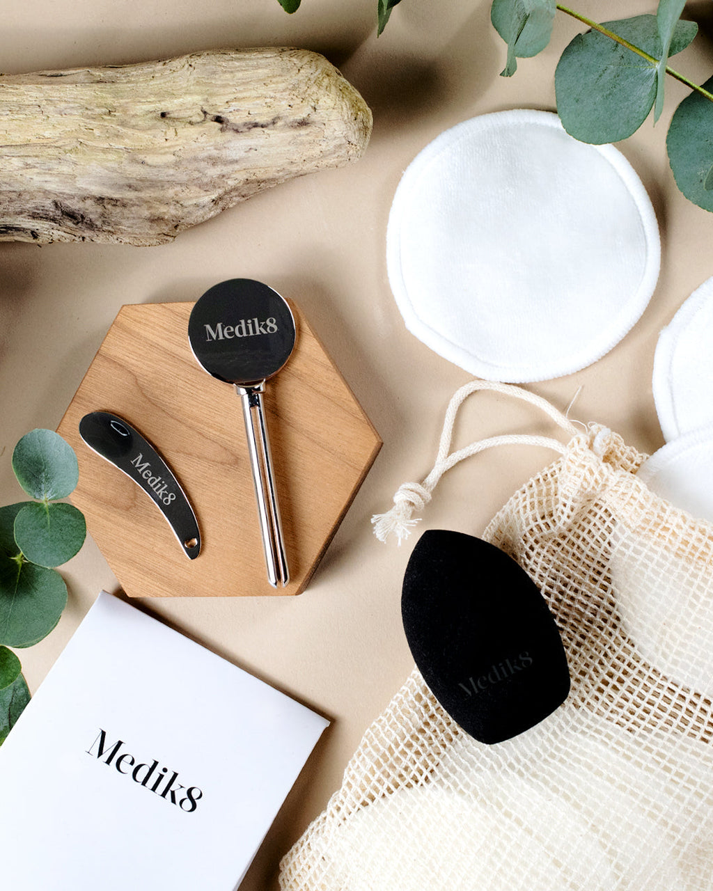 Una composición en flat lay de accesorios ecológicos de Medik8 sobre un fondo beige, incluyendo un exprimidor metálico de tubos, una espátula metálica, discos de algodón reutilizables de bambú, una bolsa de malla y una esponja de maquillaje negra.