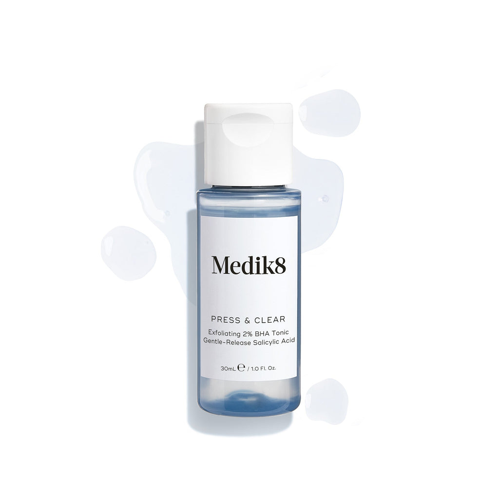 Botella de Medik8 Press & Clear (150 ml) mostrada sobre un fondo blanco con salpicaduras de tónico azul a su alrededor. El texto destaca la fórmula de tónico exfoliante con 2 % de BHA.