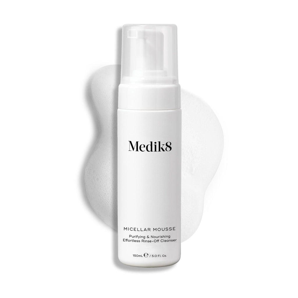 Botella con dispensador de Medik8 Micellar Mousse exhibida sobre un fondo blanco con una extensión de espuma blanca detrás, resaltando la textura del producto.
