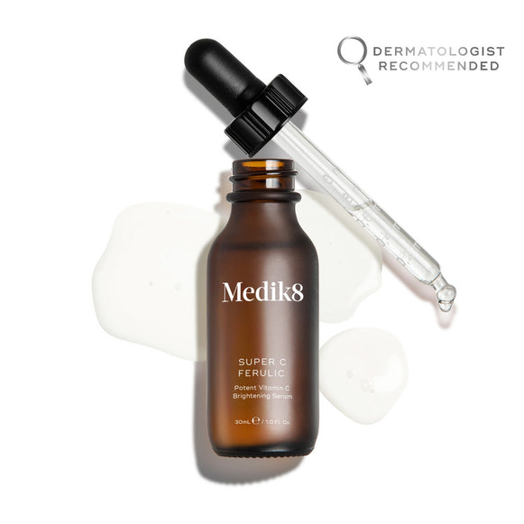 Frasco del sérum Super C Ferulic de Medik8, mostrado abierto con su pipeta de cristal apoyada en el borde, sobre un fondo blanco limpio con gotas translúcidas de sérum. El frasco de cristal ámbar lleva el logotipo de Medik8 y la etiqueta «SUPER C FERULIC Potent Vitamin C Brightening Serum» en un formato de 30 ml.-20