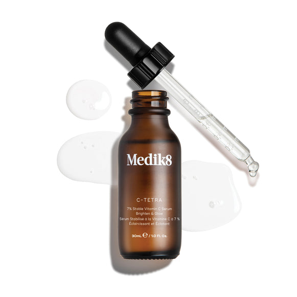 Packshot cenital de una botella de 30 mL de Medik8 C-Tetra Lipid Vitamin C Radiance Serum. La botella de vidrio ámbar está destapada y colocada en el centro, con su tapa negra con gotero apoyada diagonalmente detrás de ella. Un sérum líquido transparente está salpicado y extendido artísticamente sobre el fondo blanco limpio alrededor de la botella, con algunas gotas visibles en la pipeta de vidrio. La iluminación es brillante, proyectando sombras suaves hacia la izquierda y creando una estética clínica y el-2
