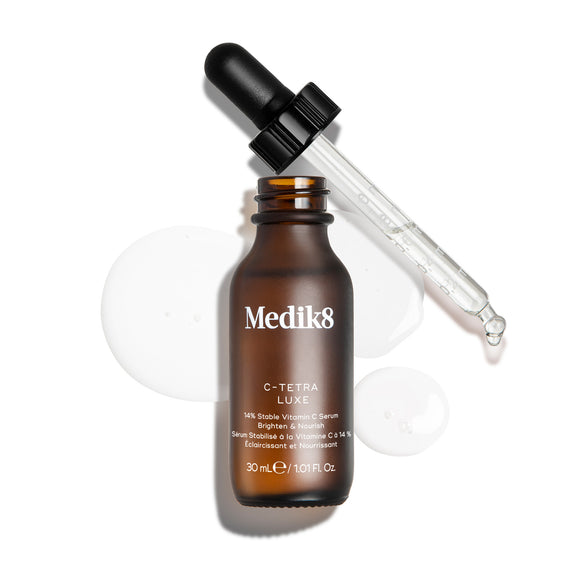 Botella de sérum Medik8 C-Tetra Luxe mostrada abierta con su pipeta de vidrio apoyada sobre el borde, presentada sobre un fondo blanco con gotas de sérum transparentes y translúcidas. La imagen incluye un sello «GET THE GLOSS BEAUTY AWARDS GOLD 2024» en la esquina superior izquierda y un emblema «RECOMENDADO POR DERMATÓLOGOS» con un icono de lupa en la esquina superior derecha.-8