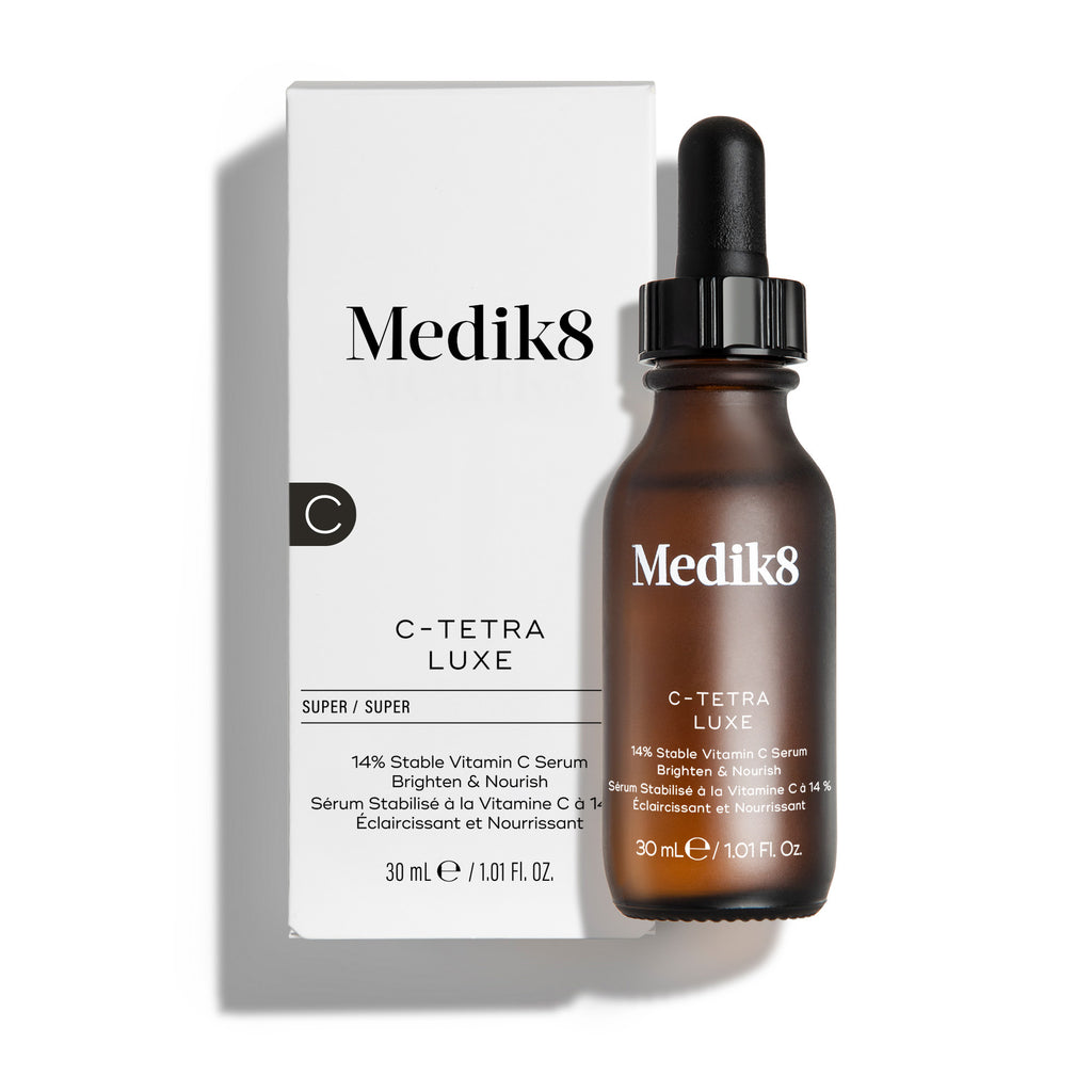 Botella de sérum Medik8 C-Tetra Luxe y su caja de cartón blanca mostradas sobre un fondo blanco limpio con sombras suaves. La botella de vidrio ámbar y la caja blanca presentan el logotipo de Medik8 e identifican el producto como «C-TETRA LUXE Lipid Vitamin C Enhanced Radiance Serum» en formato de 30 ml.