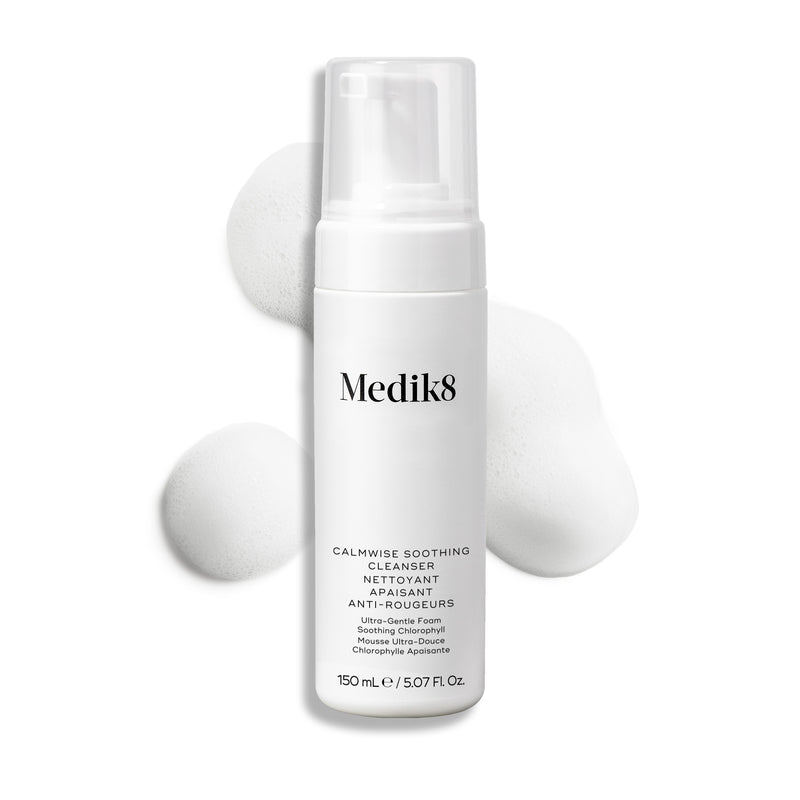 Botella con dispensador de Medik8 Calmwise Soothing Cleanser mostrada sobre un fondo blanco con una capa de espuma blanca detrás, destacando la textura del producto.