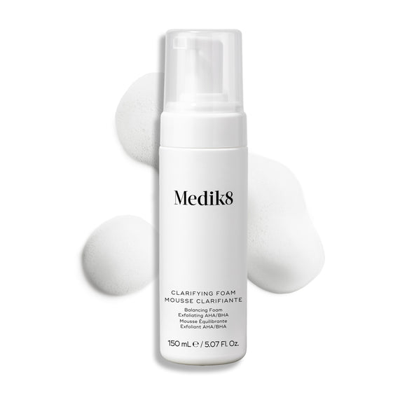 Botella con dispensador de Medik8 Clarifying Foam exhibida sobre un fondo blanco limpio junto a tres porciones de espuma blanca y ligera. El texto identifica el producto como un limpiador refrescante para imperfecciones con AHA/BHA de 150 mL.-33