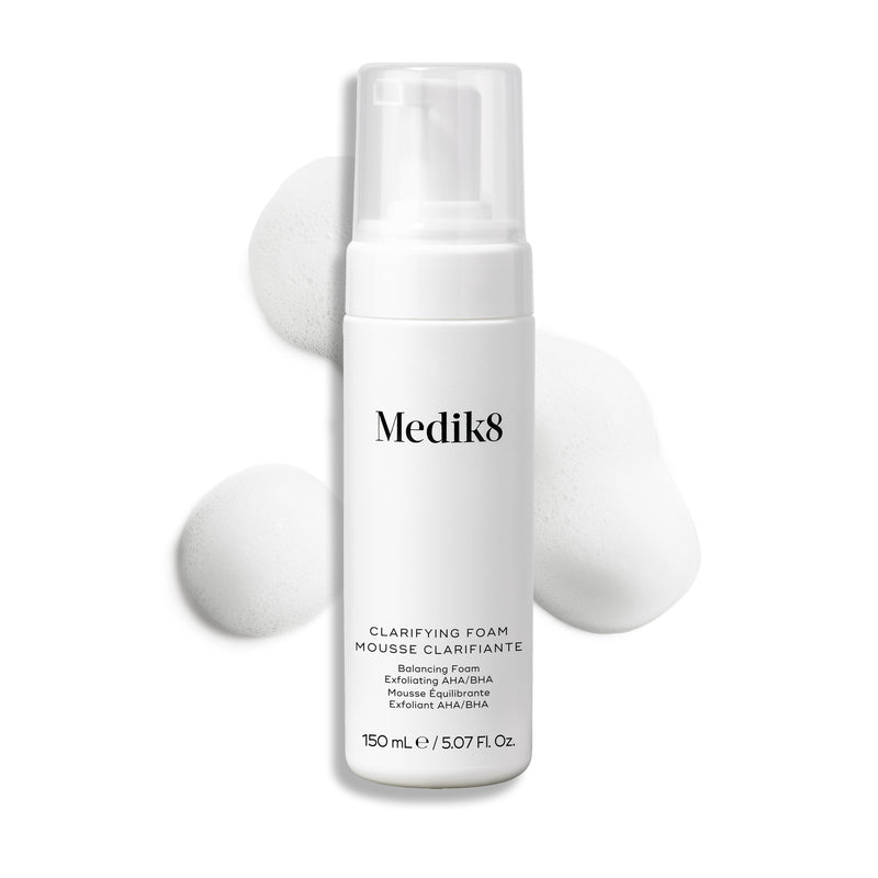 Botella con dispensador de Medik8 Clarifying Foam exhibida sobre un fondo blanco limpio junto a tres porciones de espuma blanca y ligera. El texto identifica el producto como un limpiador refrescante para imperfecciones con AHA/BHA de 150 mL.