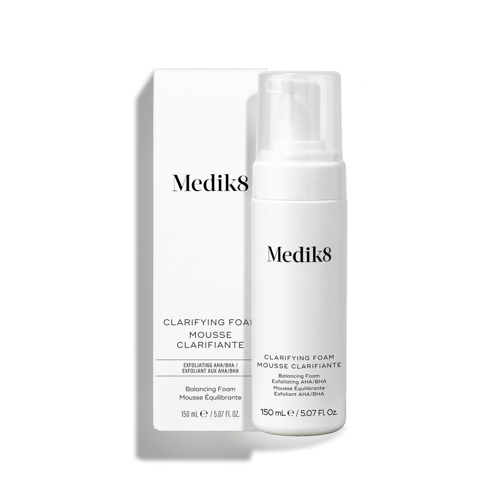 La botella con dispensador de Medik8 Clarifying Foam se encuentra junto a su caja exterior blanca a juego sobre un fondo blanco limpio. El texto en ambos identifica el producto como un limpiador refrescante para imperfecciones con AHA/BHA de 150 mL.