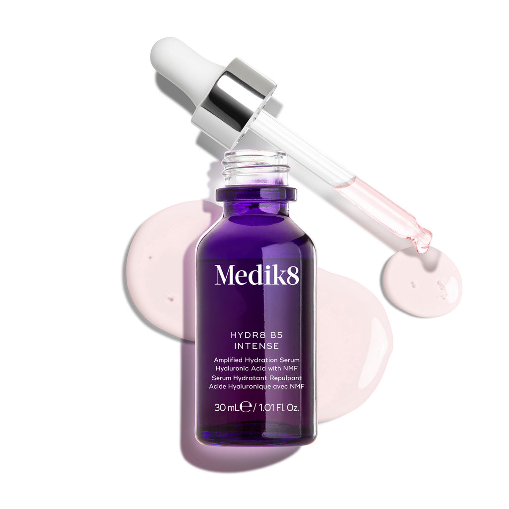 Un frasco cuentagotas de vidrio de Medik8 Hydr8 B5 Intense en color púrpura intenso, mostrado con una pipeta abierta colocada sobre gotas de sérum rosa claro sobre un fondo blanco limpio. El texto destaca la fórmula como ácido hialurónico con NMF (factores naturales de hidratación) para reponer y potenciar, en tamaño de 30 ml.