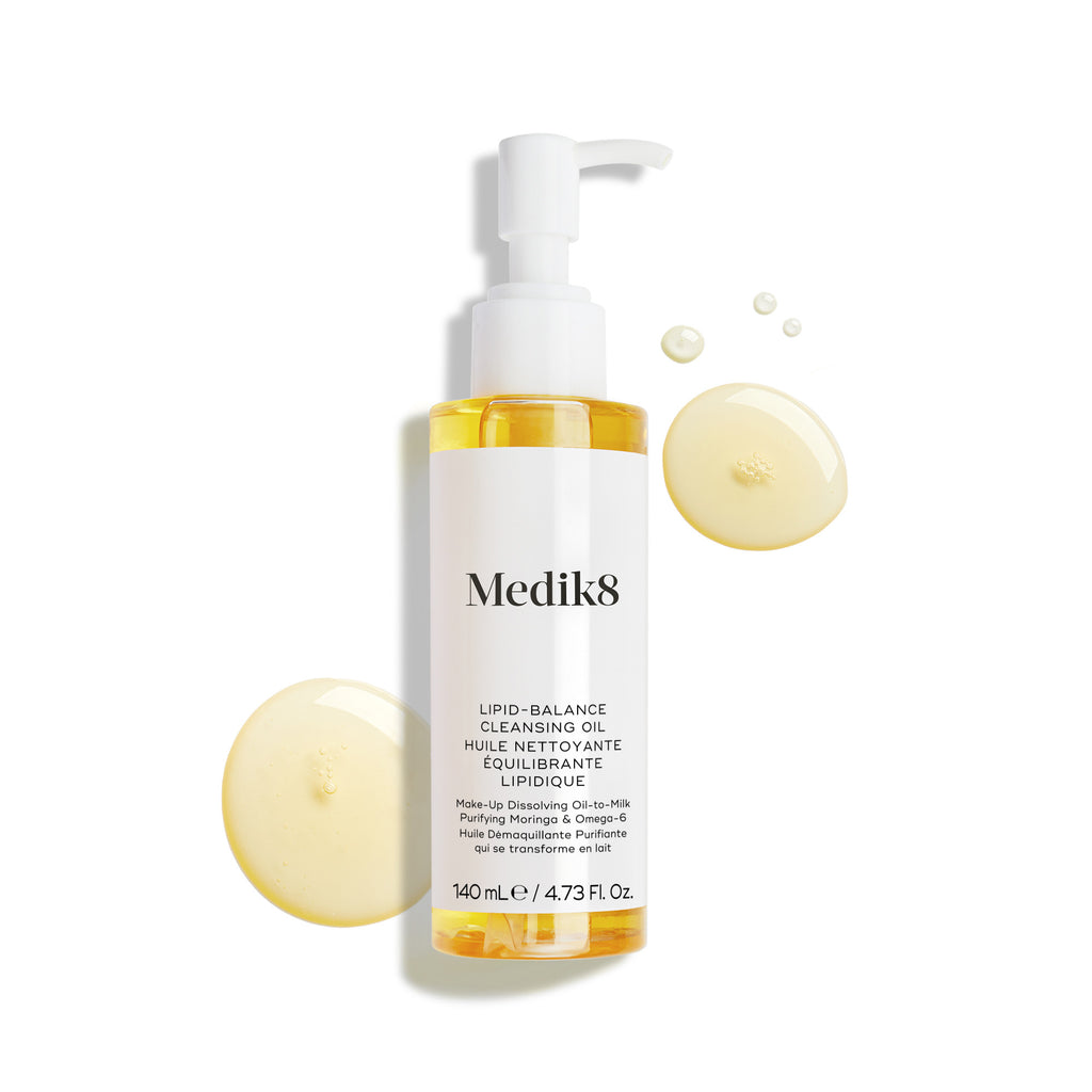 La botella de Medik8 Lipid-Balance Cleansing Oil se encuentra sobre un fondo blanco. Gotas de aceite dorado rodean la botella, resaltando su textura rica y fluida.