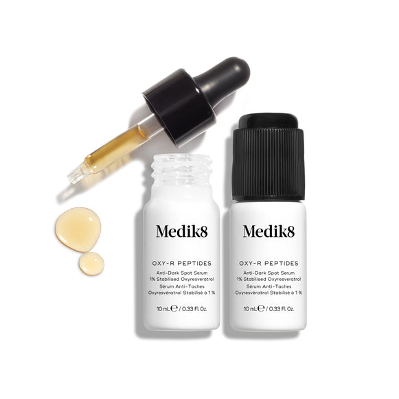 Una caja de cartón blanca de Medik8 Oxy-R Peptides está colocada junto a dos pequeños frascos blancos del sérum. Uno de los frascos tiene un cuentagotas negro y el otro un tapón negro.-37