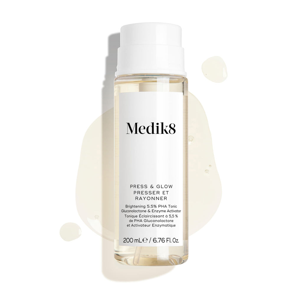 Botella del tónico Medik8 Press & Glow sobre un fondo blanco limpio con gotas translúcidas de líquido y un pequeño charco, destacando su textura acuosa. El texto identifica el producto como un tónico exfoliante diario con PHA y activador enzimático de 200 mL.