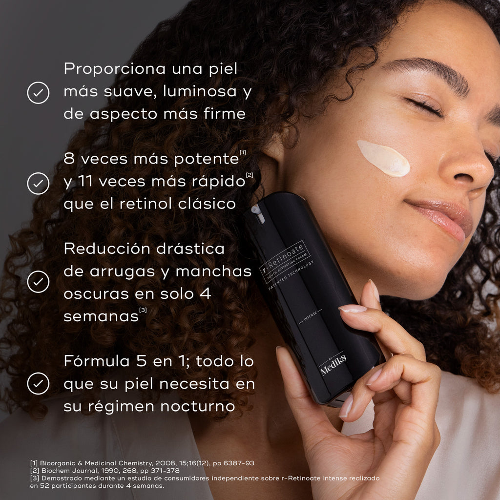 Una persona con cabello rizado sostiene un frasco de Medik8 r-Retinoate Intense Youth Activating Cream junto a su rostro, que tiene una pequeña muestra de la crema color melocotón en la mejilla.