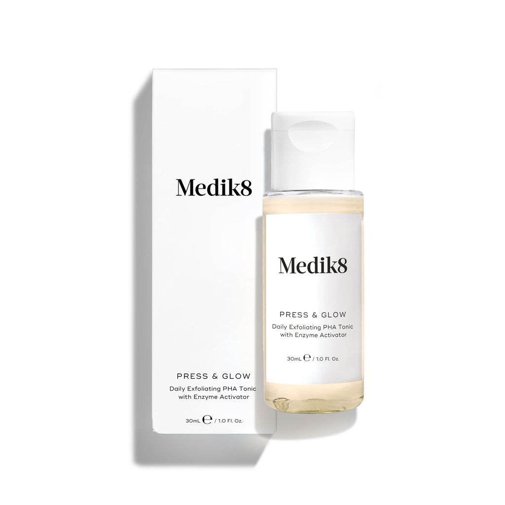 Botella de tónico Medik8 Press & Glow colocada delante de su caja de cartón blanca a juego sobre un fondo blanco limpio. El texto en ambos identifica el producto como formato de viaje de 30 ml: «Tónico exfoliante diario con PHA y activador enzimático».
