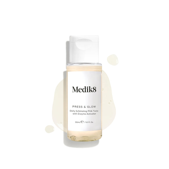 Botella transparente de viaje de 30 ml de Medik8 Press & Glow tónico con tapón blanco, mostrada sobre un fondo blanco limpio. La botella descansa sobre un charco del líquido amarillo pálido con gotas dispersas a su alrededor, destacando su textura ligera. El texto de la etiqueta dice: «Tónico exfoliante diario con PHA y activador enzimático».
-38