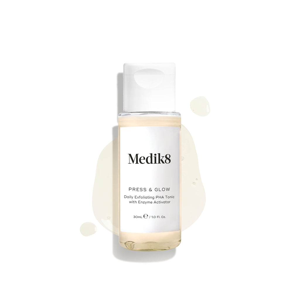 Botella transparente de viaje de 30 ml de Medik8 Press & Glow tónico con tapón blanco, mostrada sobre un fondo blanco limpio. La botella descansa sobre un charco del líquido amarillo pálido con gotas dispersas a su alrededor, destacando su textura ligera. El texto de la etiqueta dice: «Tónico exfoliante diario con PHA y activador enzimático».
