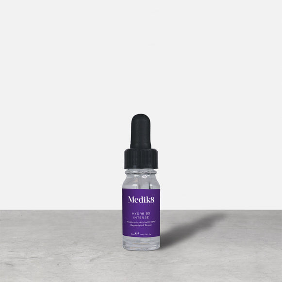 Botella de vidrio con gotero de 8 ml de Medik8 HYDR8 B5 INTENSE en color púrpura colocada junto a su caja de embalaje blanca sobre una superficie gris con fondo blanco. El texto en ambos indica: «Medik8, HYDR8 B5 INTENSE, Ácido hialurónico con NMF Replenish & Boost, 8 ml / 0,27 fl. oz.».
-37