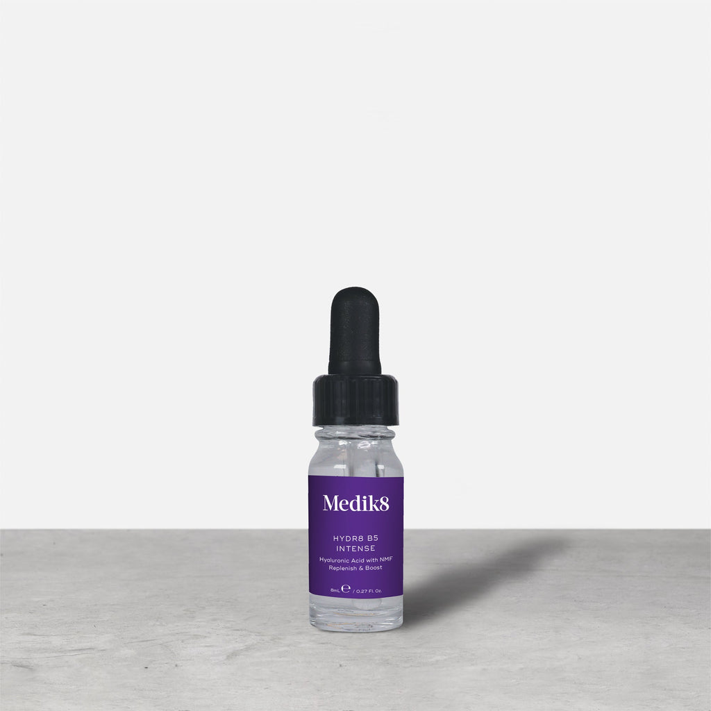 Botella de vidrio con gotero de 8 ml de Medik8 HYDR8 B5 INTENSE en color púrpura colocada junto a su caja de embalaje blanca sobre una superficie gris con fondo blanco. El texto en ambos indica: «Medik8, HYDR8 B5 INTENSE, Ácido hialurónico con NMF Replenish & Boost, 8 ml / 0,27 fl. oz.».
