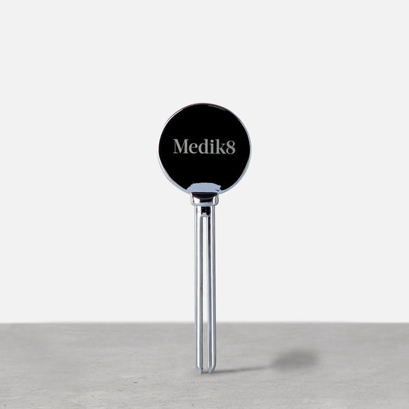 Un exprimidor metálico plateado de tubos de Medik8 en posición vertical sobre una superficie gris. El cabezal circular presenta el logotipo de Medik8, diseñado para ayudar a dispensar el producto de forma eficiente.-45