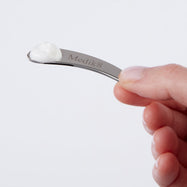 Primer plano de una mano sosteniendo una espátula metálica con la marca Medik8 y una pequeña cantidad de crema blanca en la punta, demostrando la cantidad correcta a utilizar.